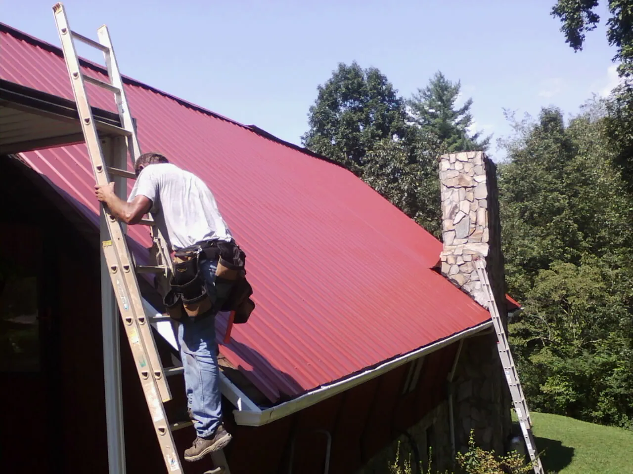 Roofer on a ladder beside a red metal roof and stone chimney — Soffit & Fascia Repair by Roofing Brothers in Collins
